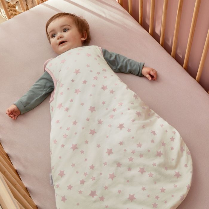 A happy baby lay in a cot wearing the Silentnight Safe Nights Stars Baby Sleep Bag with pink stars